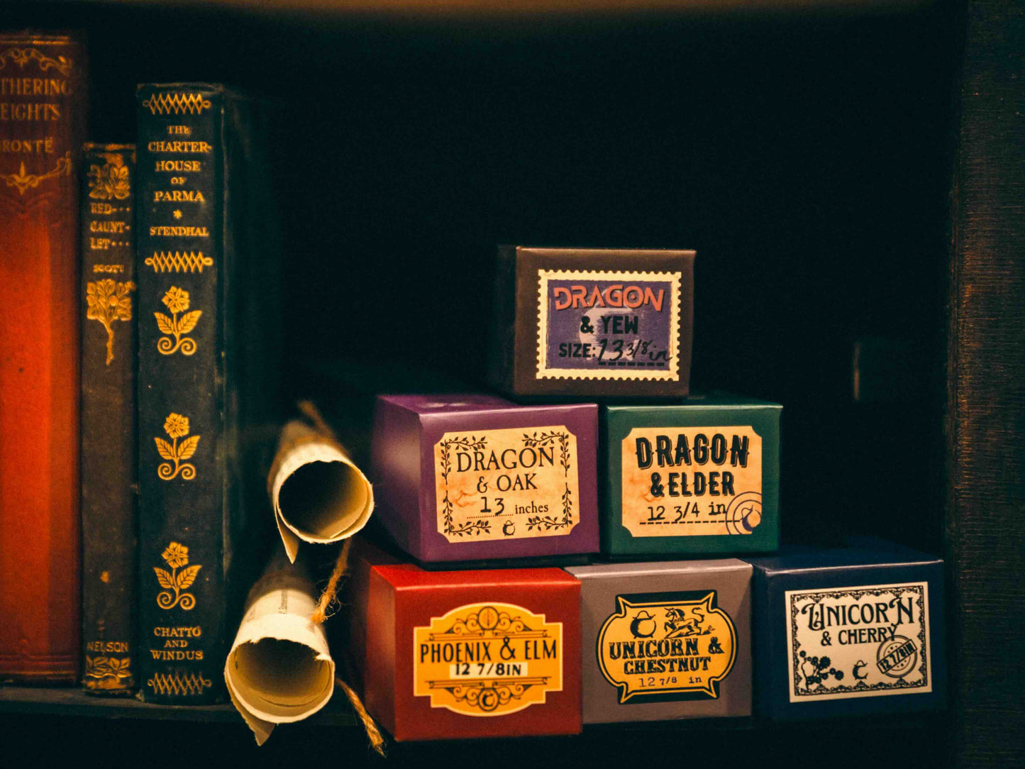 Six boxed wands labeled with fantasy themes, including Dragon, Phoenix, and Unicorn, in a magical display on a shelf of old books at The Cauldron.