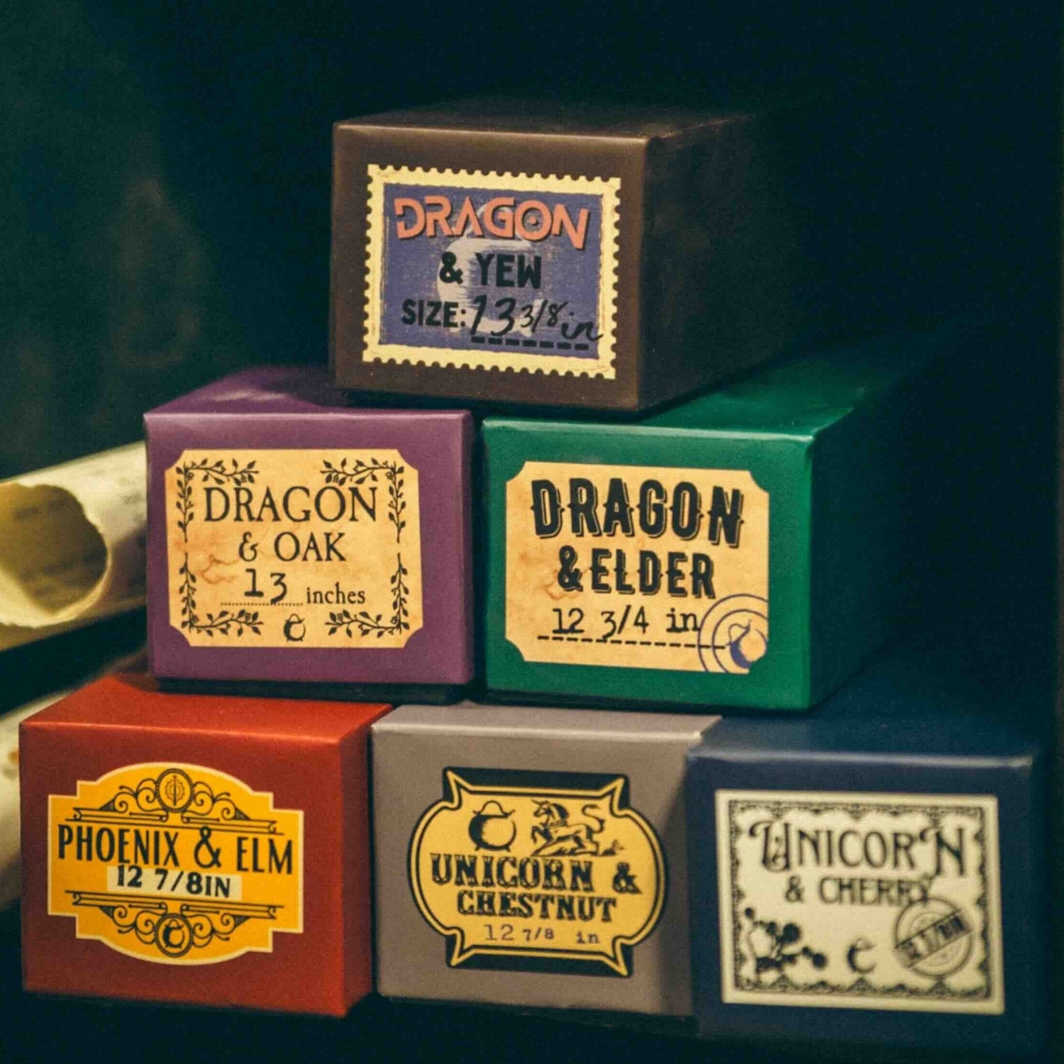 Stack of six magical wand boxes labeled for Dragon & Oak, Dragon & Yew, Dragon & Elder, Phoenix & Elm, Unicorn & Chestnut, and Unicorn & Cherry wands, displayed on a bookshelf with vintage books – crafted by The Cauldron.